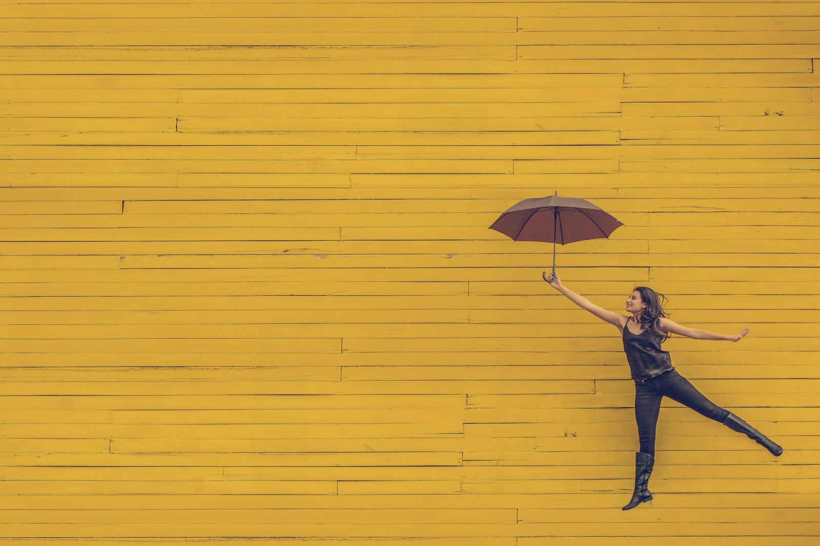 Frau springt mit Regenschirm vor einer gelben Holzwand